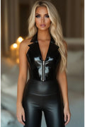 A blonde woman wearing a shiny black sleeveless bodysuit with a high collar and front zipper, paired with matching black faux-leather pants, set against a softly blurred indoor background.